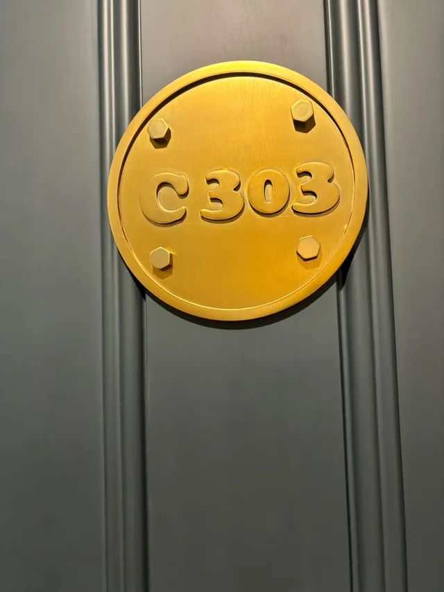 Finished brushed-gold C303 round number plate with four hex-bolt details on a dark grey door