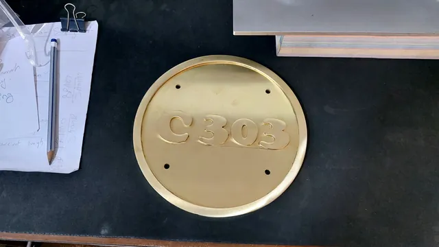 Raw brass round number plate with raised C303 letters on the workshop bench